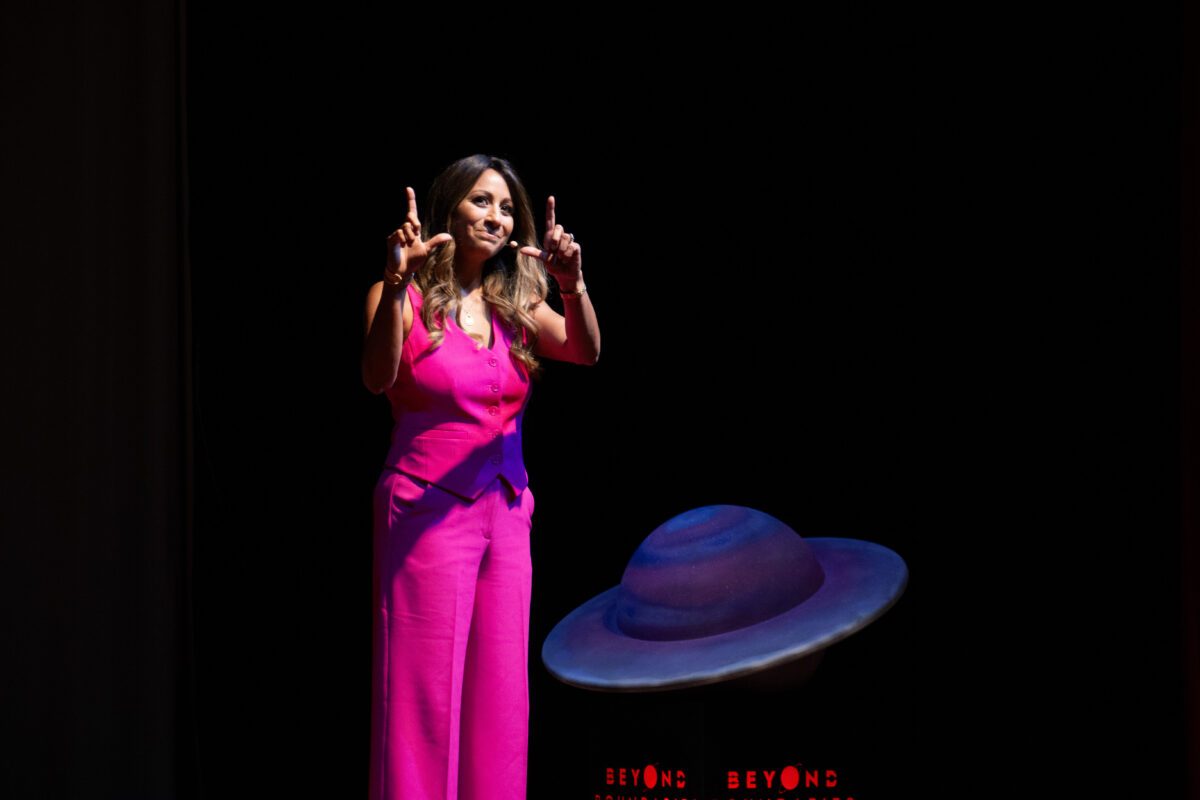 A woman in a bright pink outfit speaks onstage under a spotlight, standing beside a colorful planet-shaped prop during a live event.