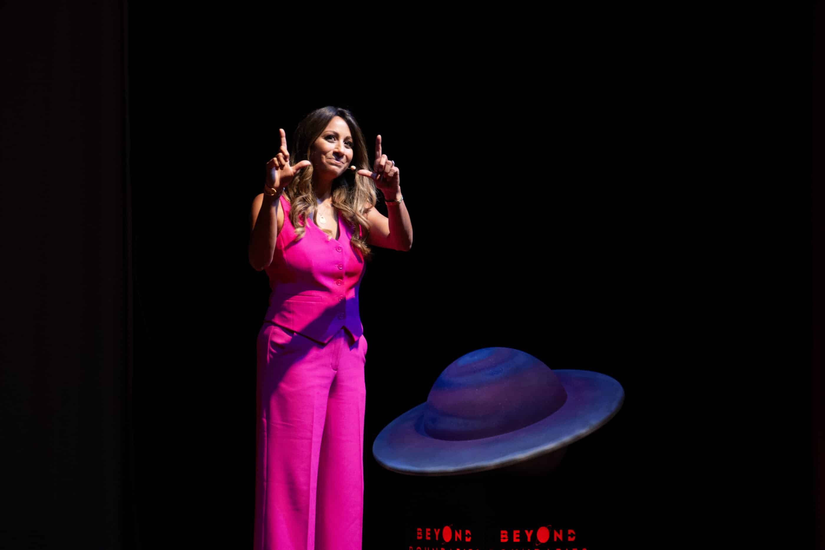A woman in a bright pink outfit speaks onstage under a spotlight, standing beside a colorful planet-shaped prop during a live event.